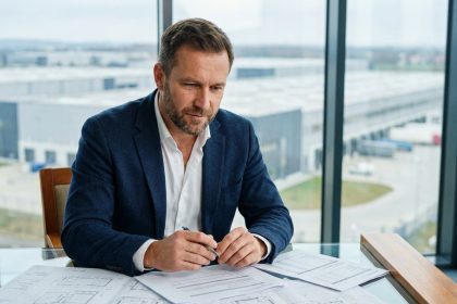 Investisseur immobilier analysant des documents financiers avec vue sur un entrepôt logistique moderne